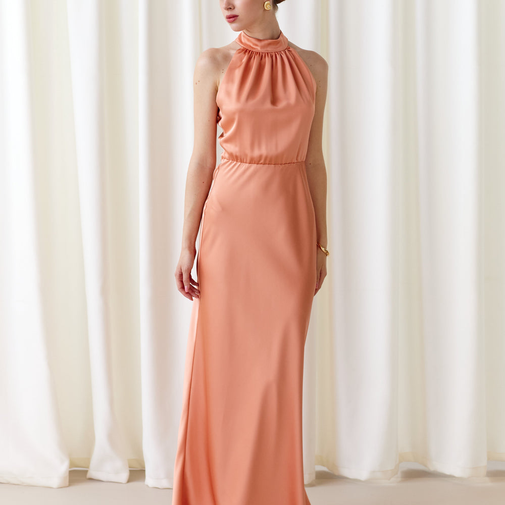 Woman wearing a coral halter neck gown standing in front of white curtains.