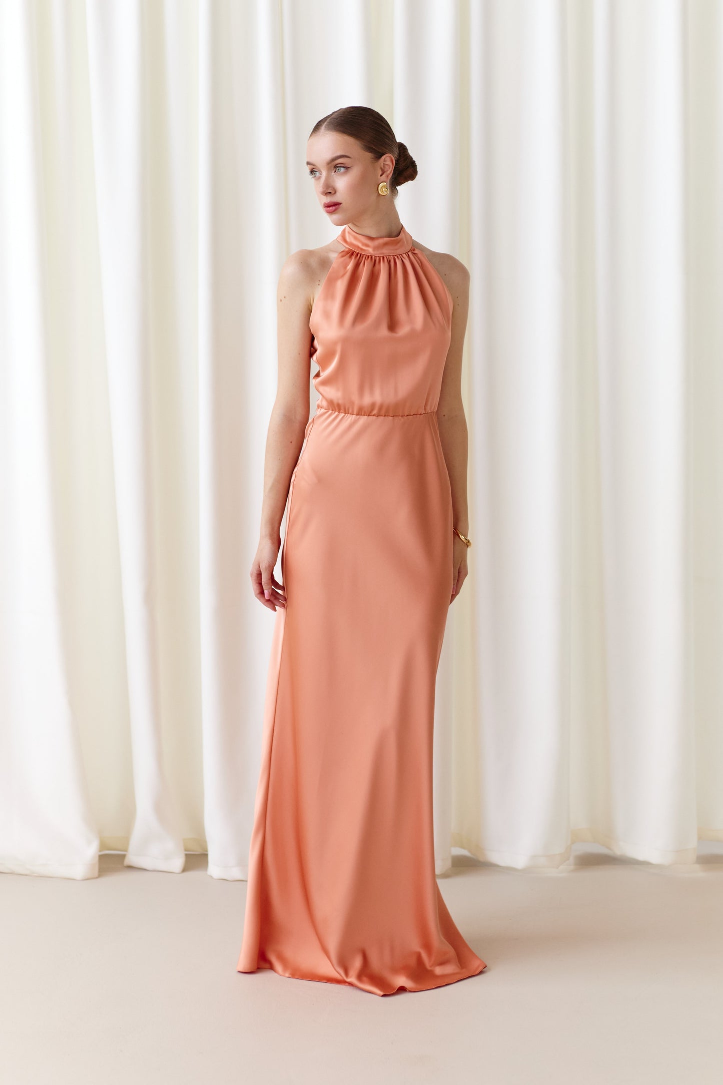 Woman wearing a coral halter neck gown standing in front of white curtains.