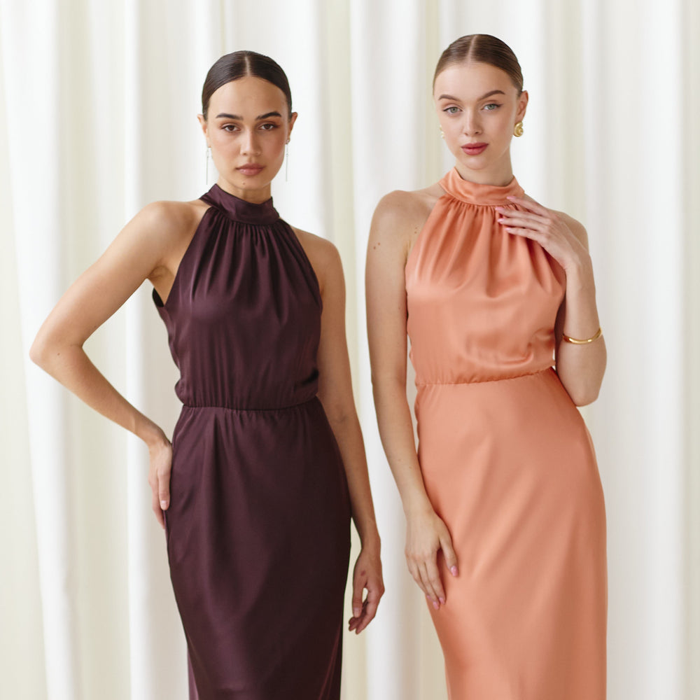 Two women wearing long, elegant dresses standing against a white curtain background.